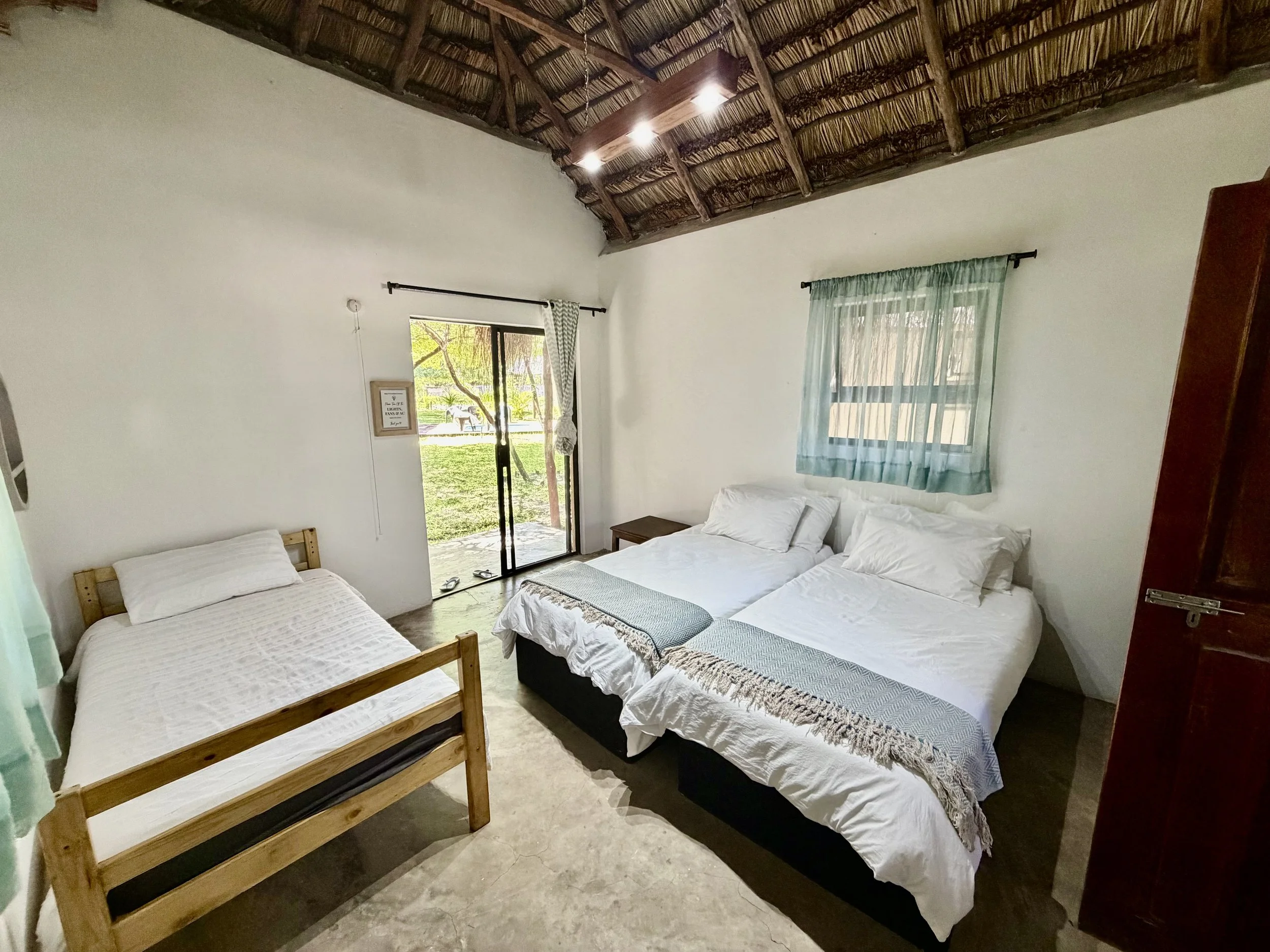 Room with white walls and beds, one with a wooden frame, and a sliding glass door leading outside, in a rustic style with a thatched roof ceiling.