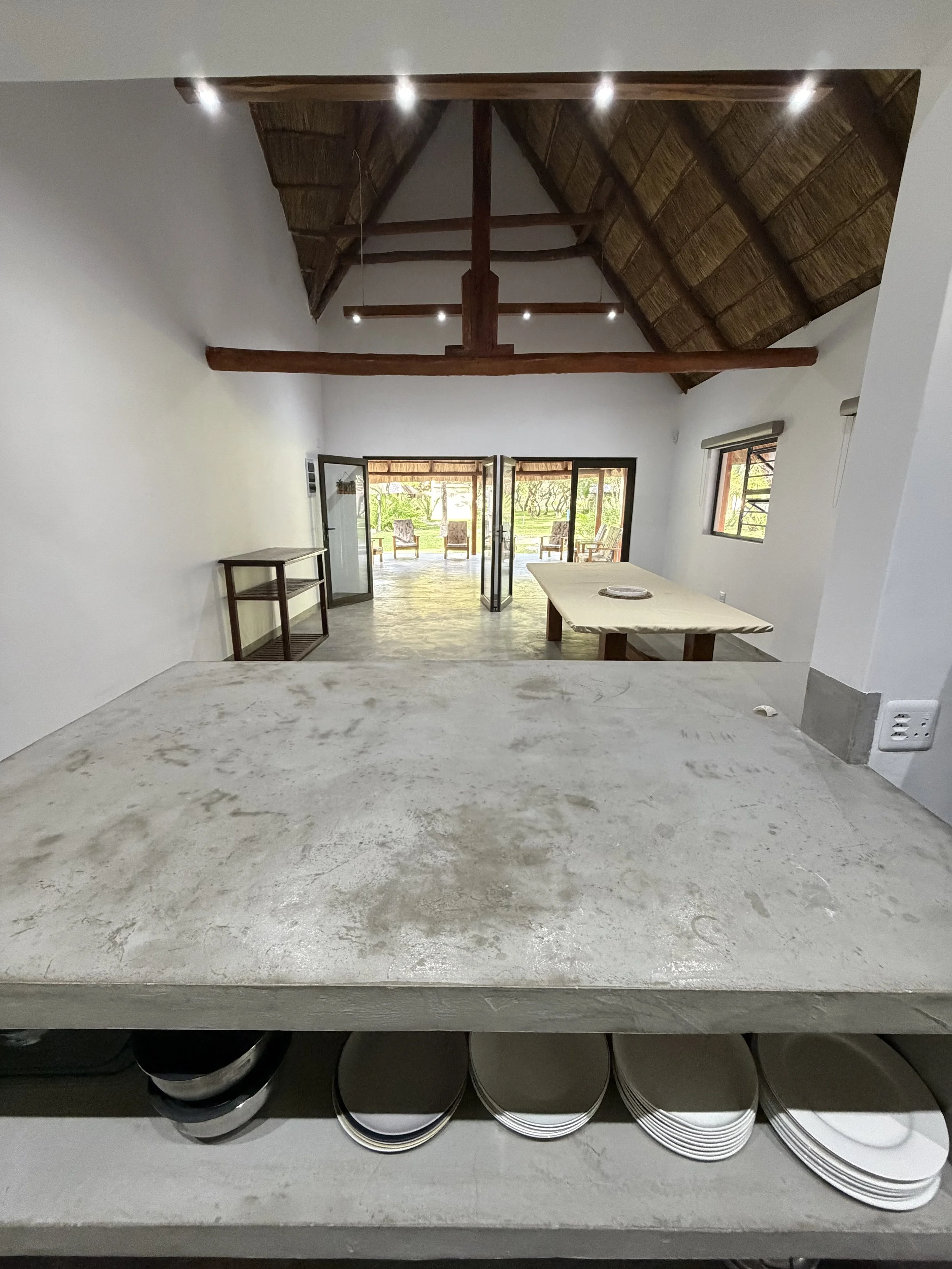 Empty room with wooden ceiling beams, open double glass doors leading outside with patio furniture, white walls, small window, and a table. The foreground shows a marble or stone countertop with a stack of plates underneath.