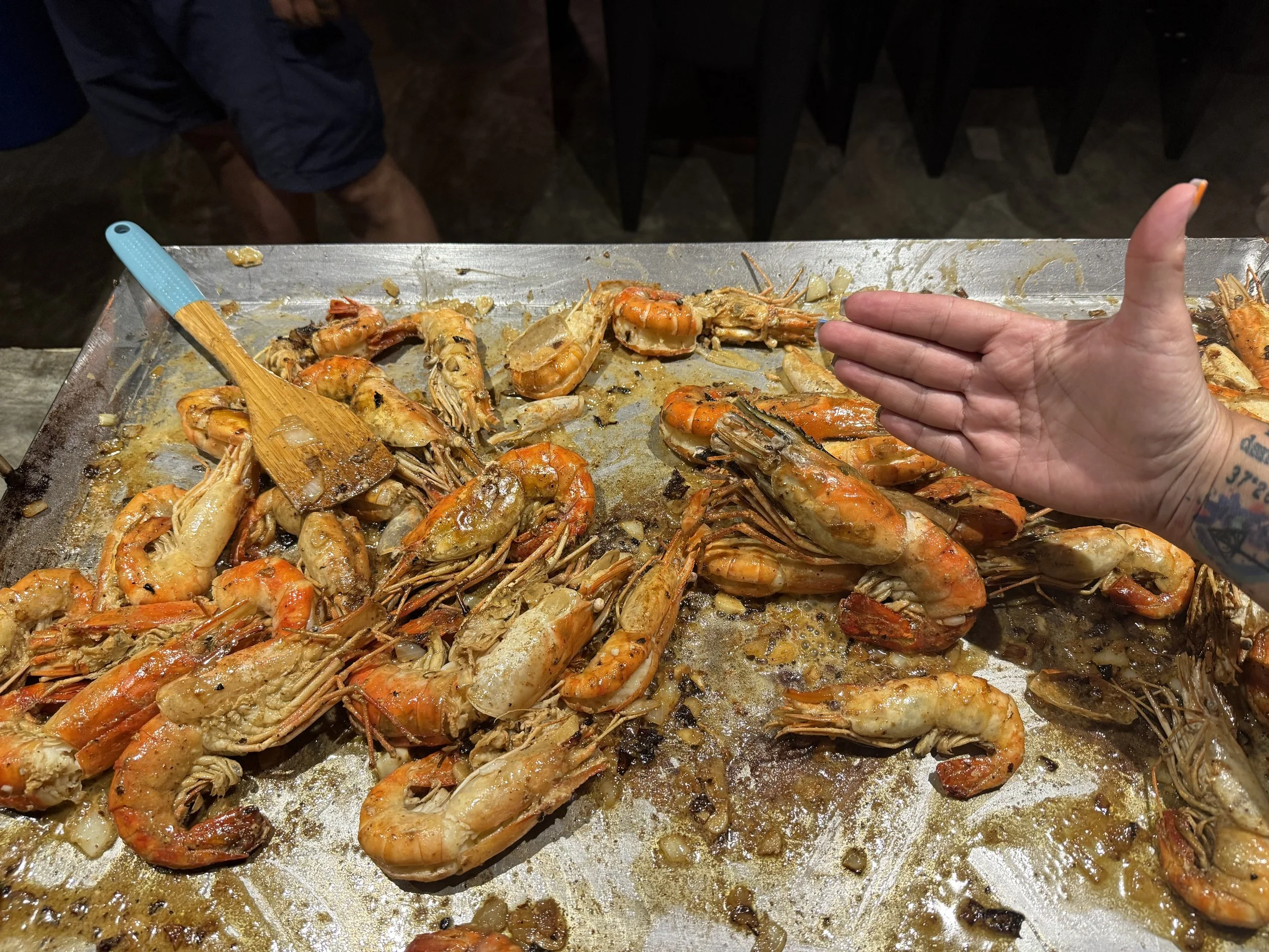 A tray of cooked shrimp with some garlic and seasoning, with a person's hand and thumb giving a thumbs up in the foreground.