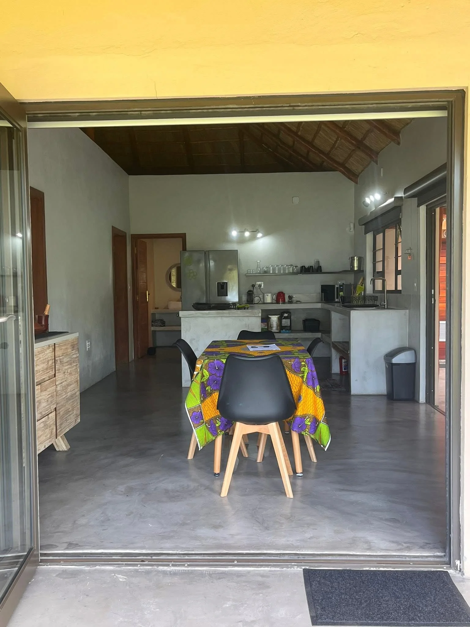 View of a kitchen with a round table covered with a colorful floral tablecloth, black chairs around it, a gray floor, and cabinets with kitchen appliances and utensils.
