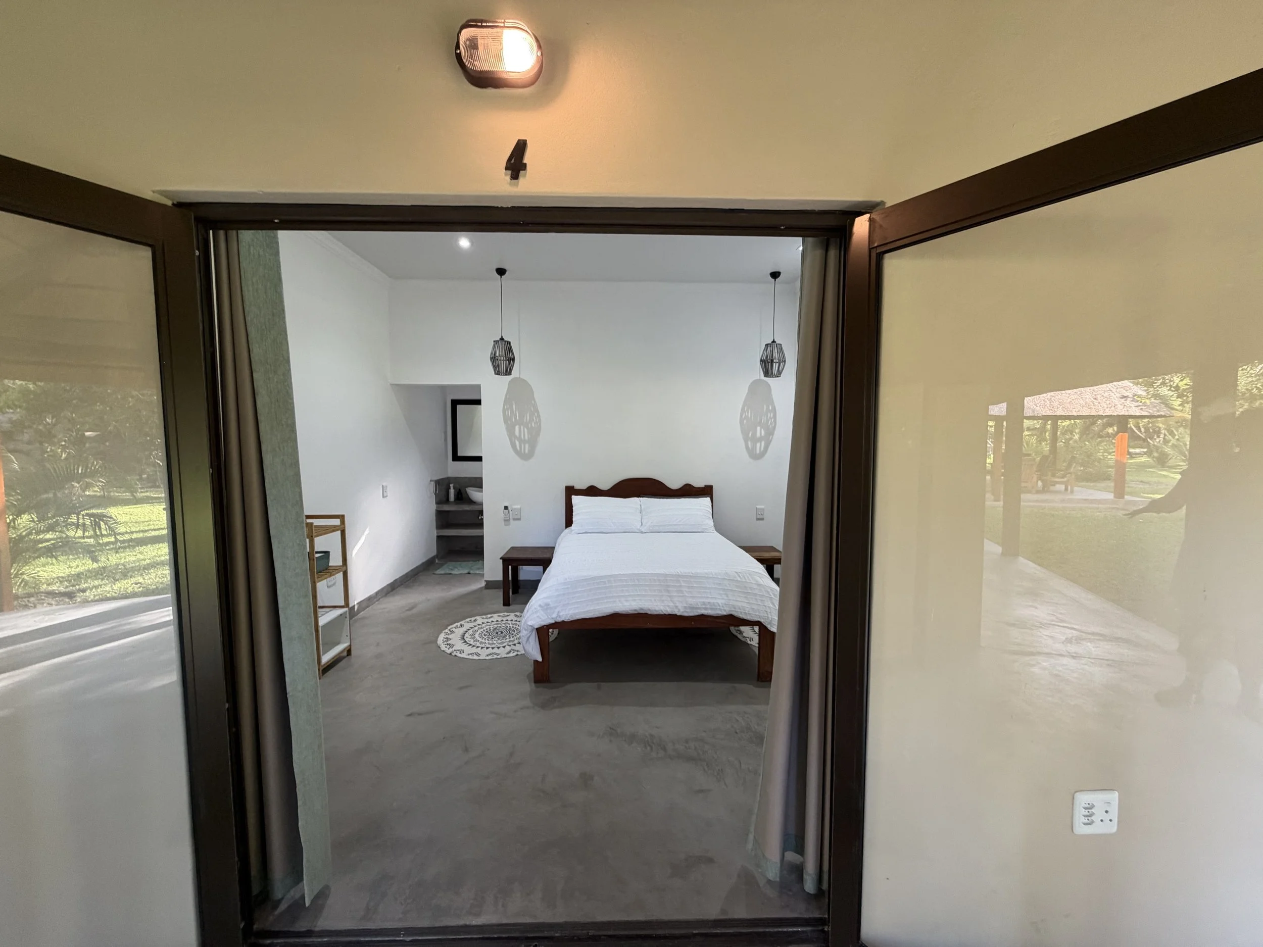 View of a bedroom with a bed, two hanging pendant lights, a small side table, a mirror, and a circular rug. The image is taken from outside the room through a glass door or window.