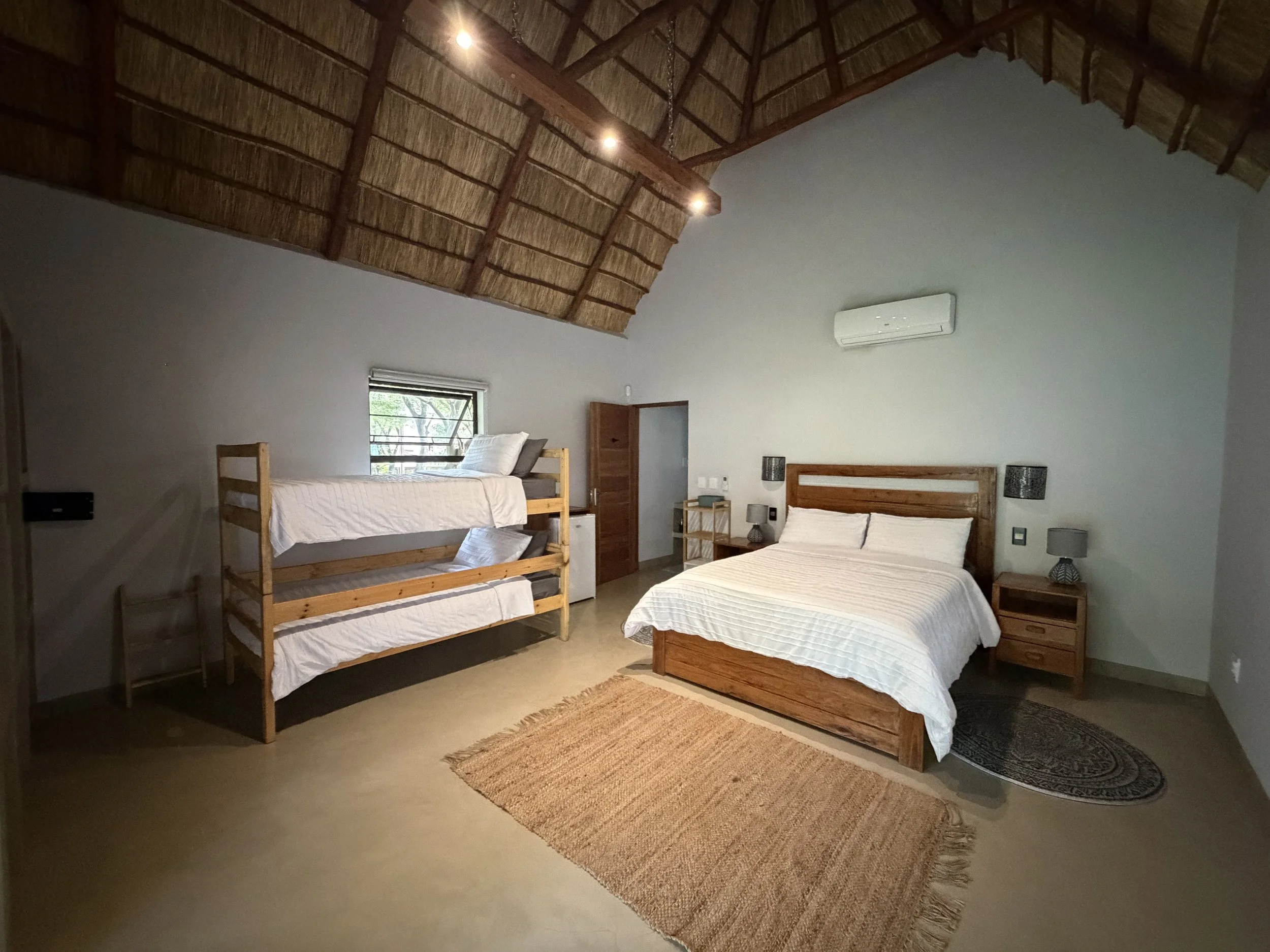 A bedroom with a high, thatched roof and two beds. One bed is a double, and the other is a bunk bed. There are side tables with lamps, a window, and an air conditioning unit on the wall.