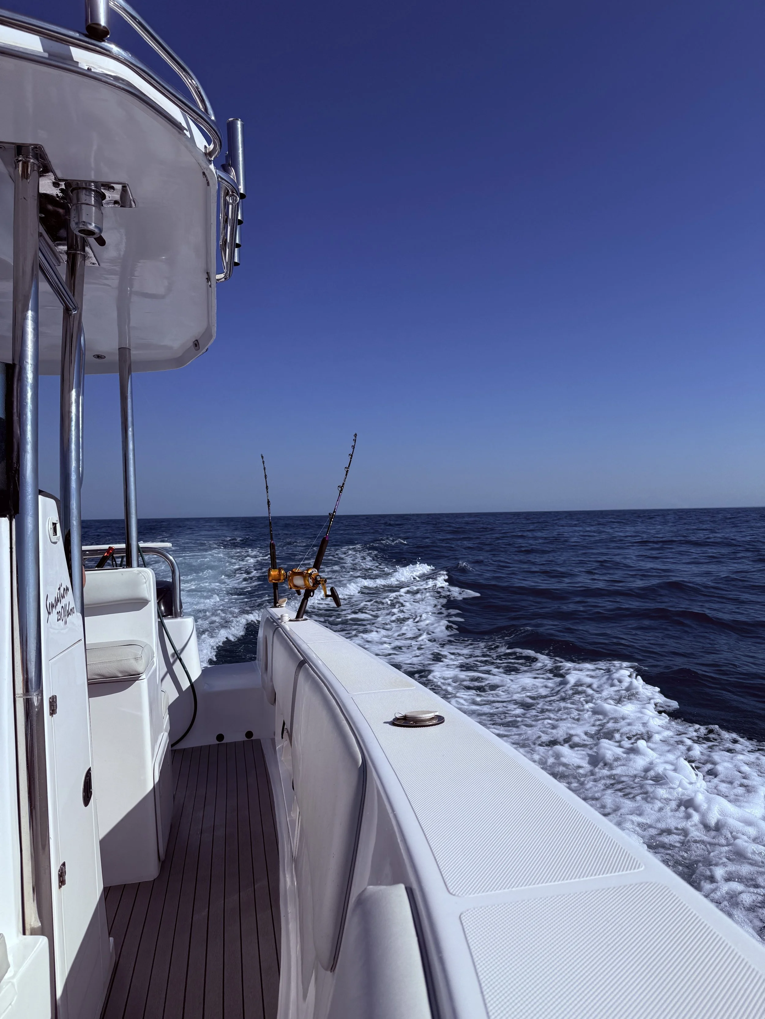 View from the side of a white boat on the open ocean, with fishing rods mounted and the wake of the boat in the water, under a clear blue sky.