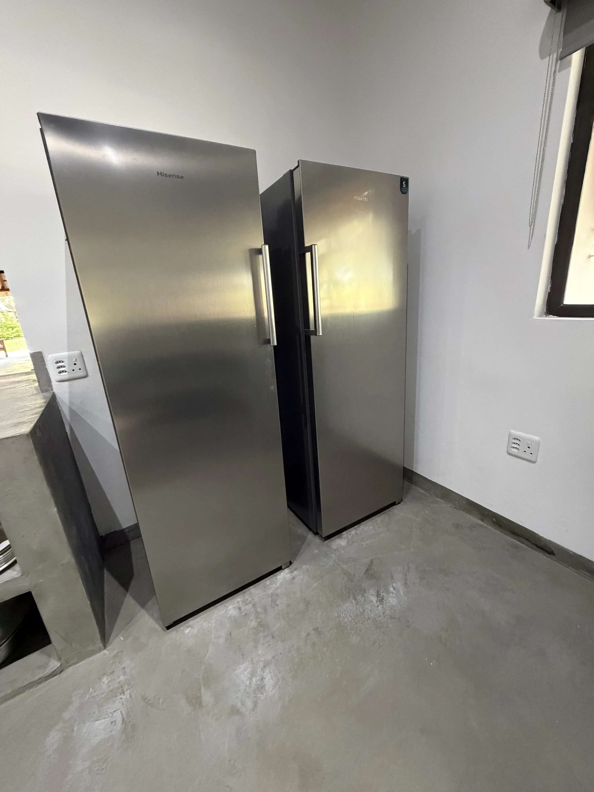 Two stainless steel refrigerators in a room with grey cement floor and white wall, window on the right, and electrical outlet on the wall.