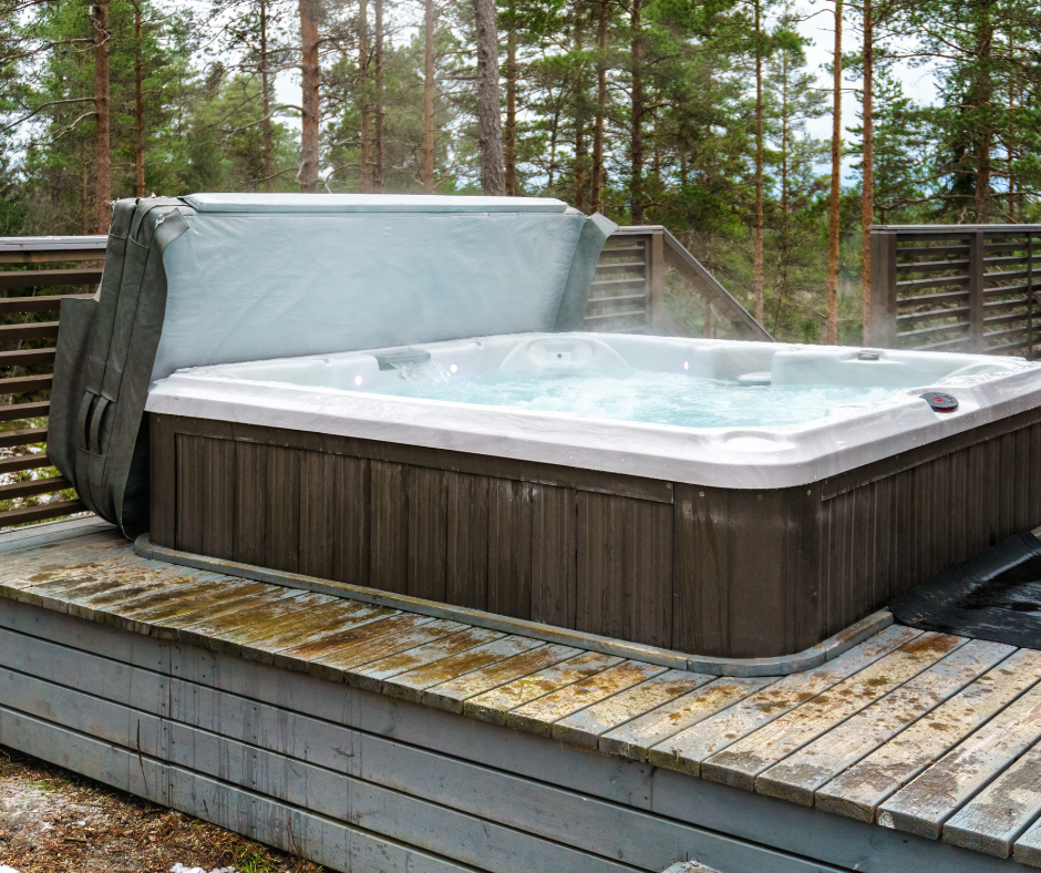 A hot tub with steam rising from the water, located on an outdoor wooden deck, surrounded by a wooden fence and trees.