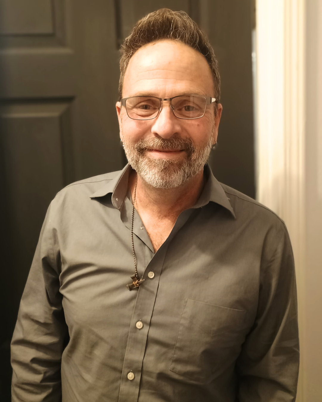A smiling man with glasses and a beard wearing a gray button-up shirt, standing indoors near a dark door and a light-colored wall.
