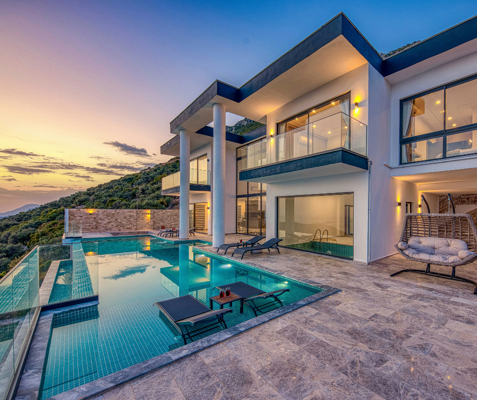 Modern house with a swimming pool, lounge chairs, and glass balconies overlooking a hillside at sunset.