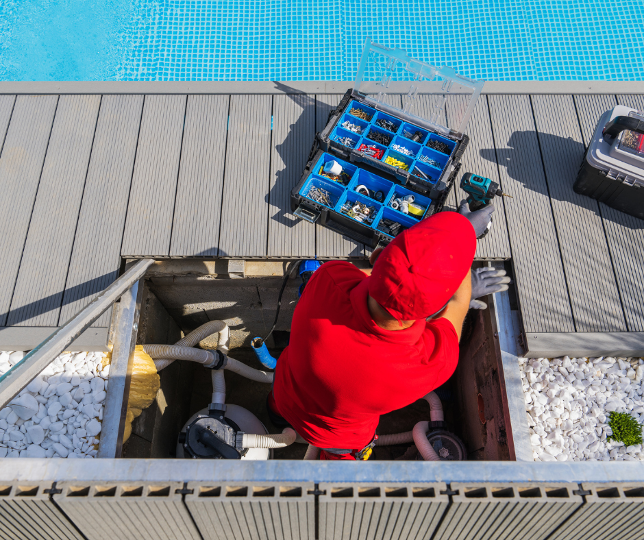 A person wearing a red shirt and red hat working on pool equipment near a swimming pool. The person is using a power drill and has a plastic tool box filled with hardware next to them.