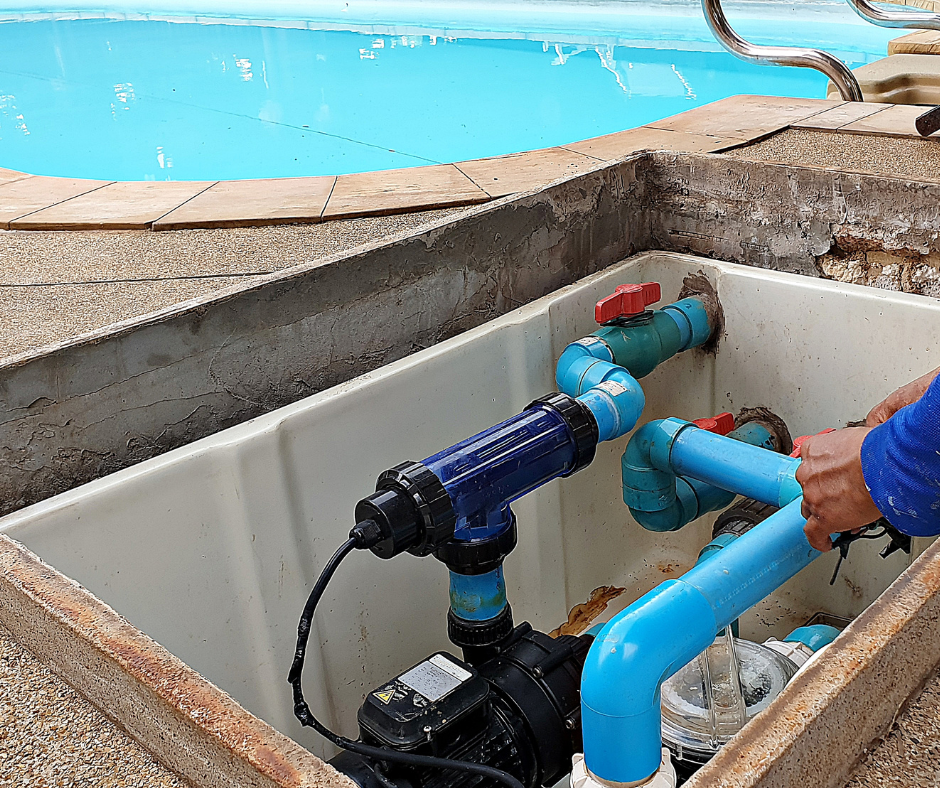 Pool equipment with blue and green pipes and a water pump, installed in a concrete basin near a swimming pool.