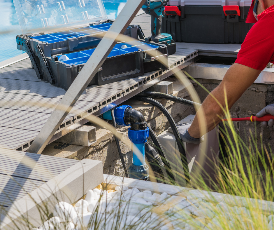 A person in a red shirt and gloves working on pool plumbing with tools near a poolside deck. There are construction materials, a blue valve, and a toolbox in the scene.