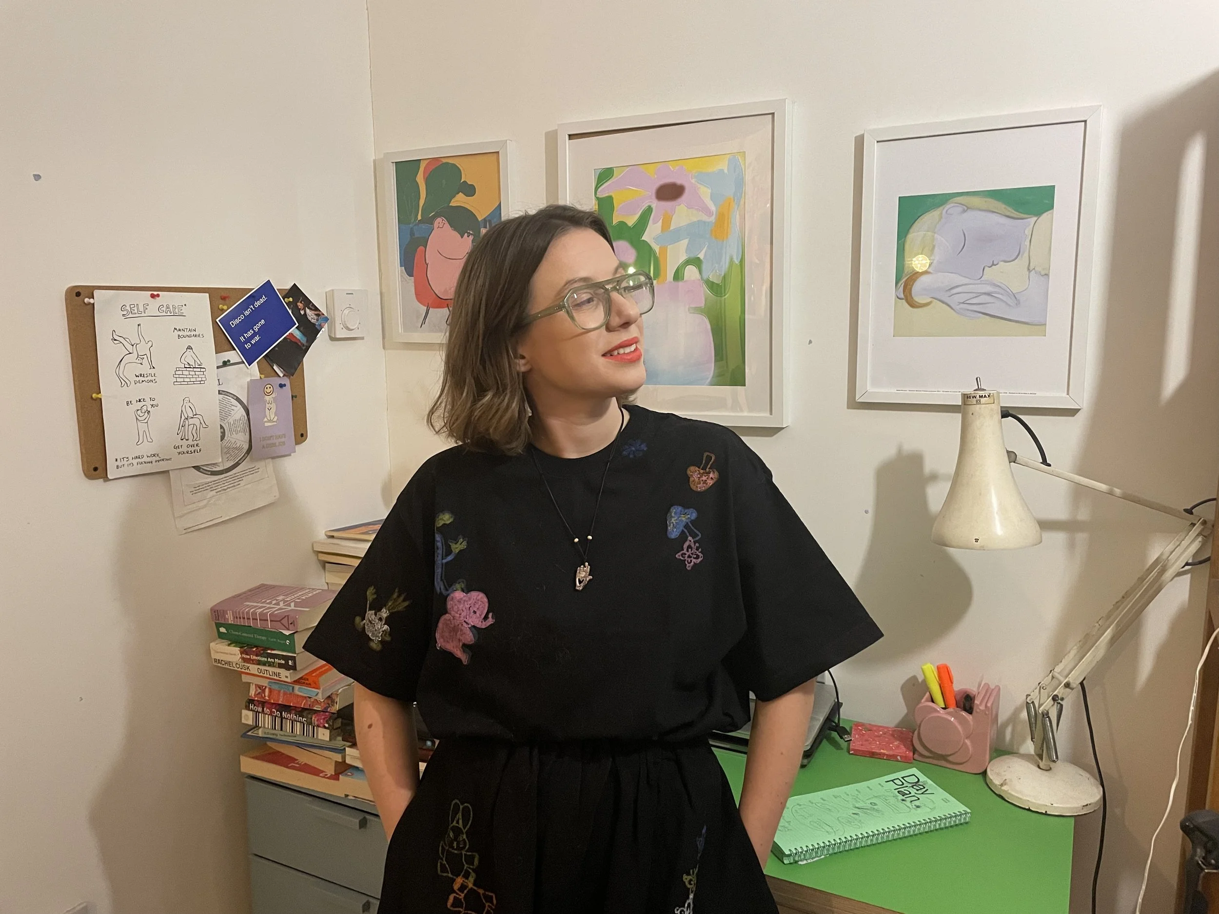 A woman with shoulder-length brown hair, glasses, and red lipstick standing in a room with colorful artwork on the wall, a desk with books, a green notebook, and a flower-shaped pen holder.