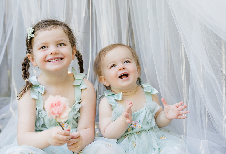 Two young girls sitting behind white sheer curtains, smiling and enjoying a moment together. The older girl has pigtails, and the younger girl is clapping and looking up with excitement. Both are wearing light-colored dresses with floral embroidery.