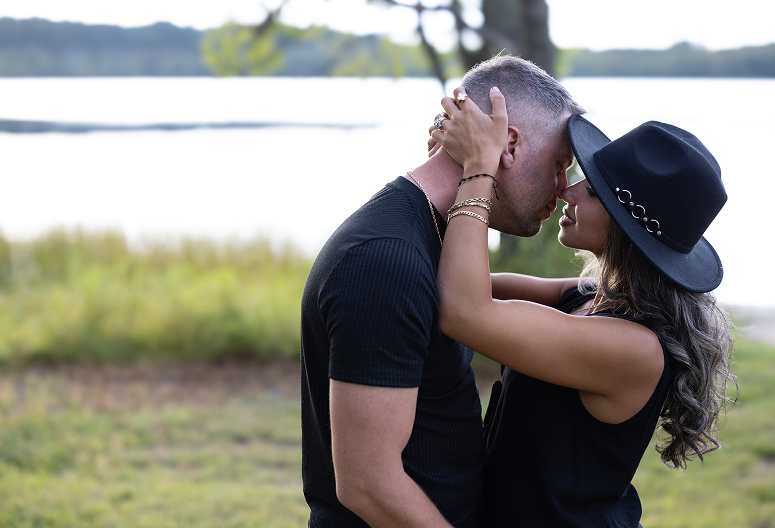 A couple is embracing and about to kiss outdoors by a lake, with the woman wearing a black hat and the man in a black shirt.
