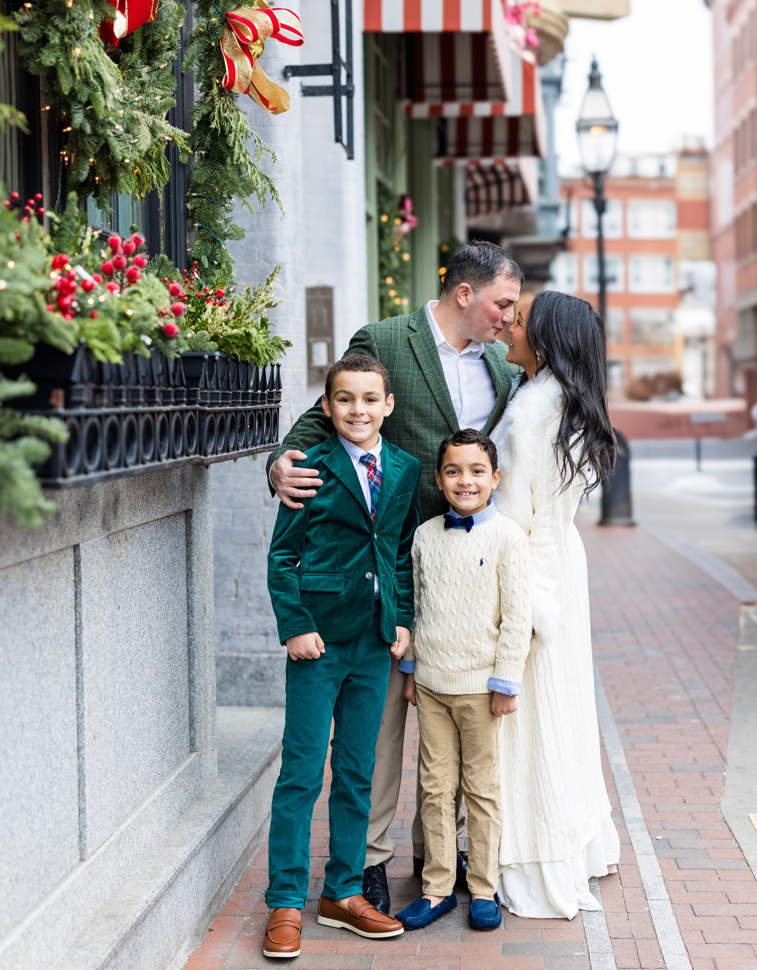 A family of four standing on a city sidewalk decorated with Christmas wreaths and red bows. The father and mother are leaning in for a kiss, while the two young sons smile at the camera.