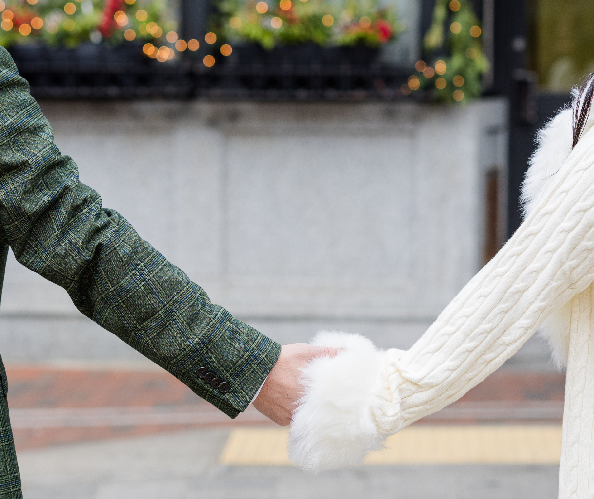 Two people holding hands, one wearing a green plaid blazer and the other a white knit sweater with fur cuffs, outdoors with blurred lights in the background.