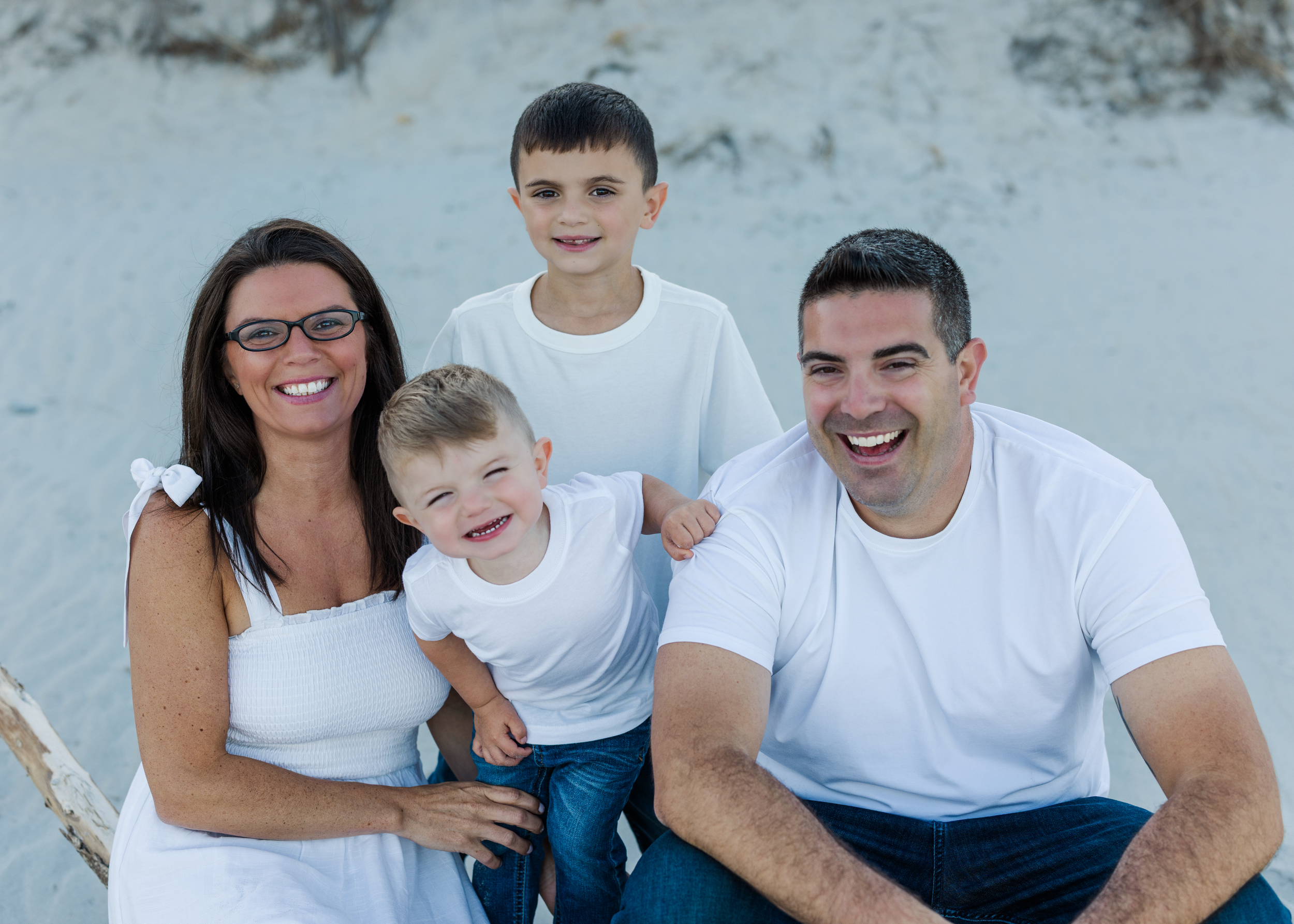A family of five smiling on a beach, mom, dad, and three young boys, all wearing white shirts.