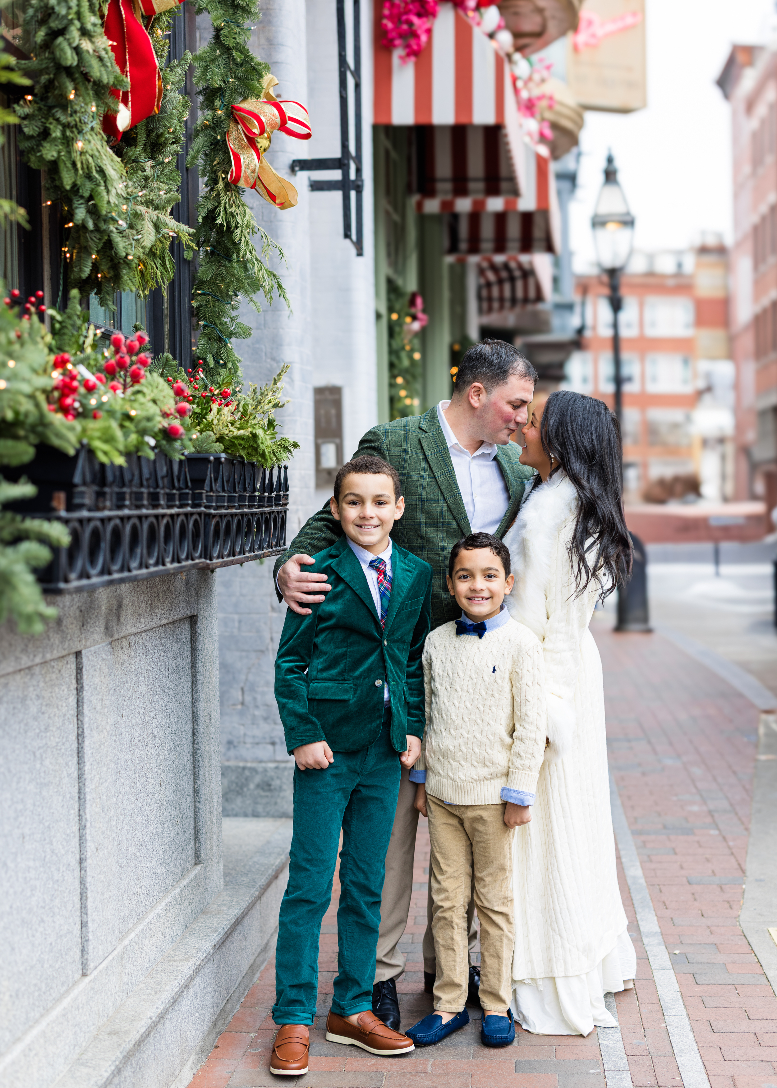 A family of four on a Christmas decorated city street, with two boys in front and a man and woman kissing in the background.