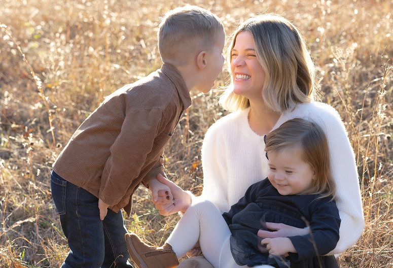 A woman with two young children outdoors in a sunny, grassy field, one boy leaning in to kiss the woman's cheek, who is smiling, while the girl sits on her lap smiling.
