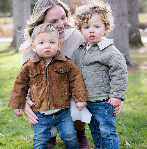A smiling woman with blonde hair holding two young boys outside in a park, both children wearing jackets and jeans.
