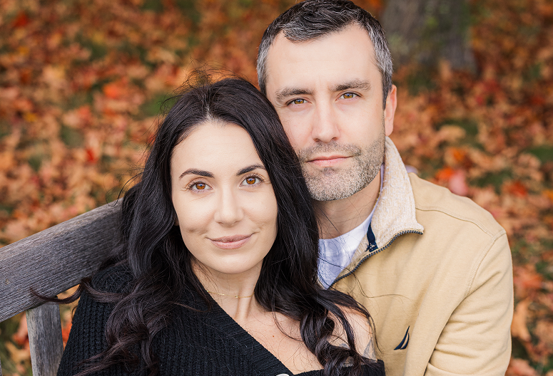 A couple sitting on a bench outdoors during autumn, surrounded by fallen leaves.