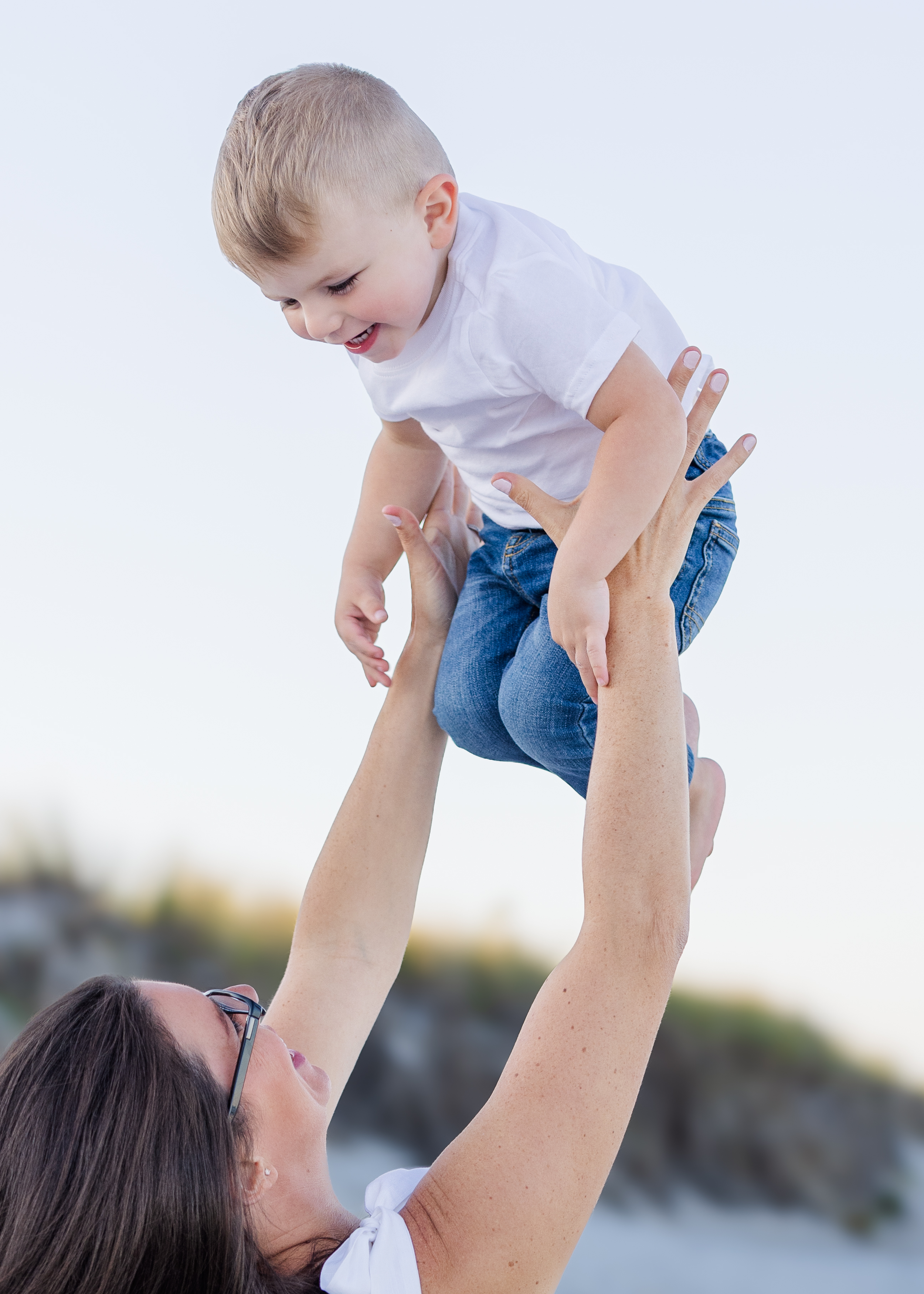A woman lifting a young boy in the air outdoors with a blurred background, possibly near a body of water or a park.