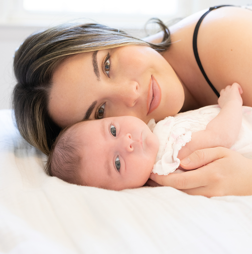 A woman lying down next to a baby, both on a bed. The woman has long brown hair and is wearing a black strap top. The baby is lying on its side with blue eyes and wearing a white outfit. They are close, looking content and relaxed.