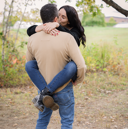 A man holding a woman in his arms outside in a park or yard with trees and grass, during fall.