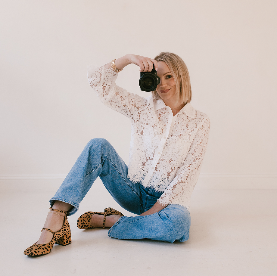 Woman with blonde hair in a lace white blouse and blue jeans sitting on the floor, taking a selfie with a camera, against a plain white background.