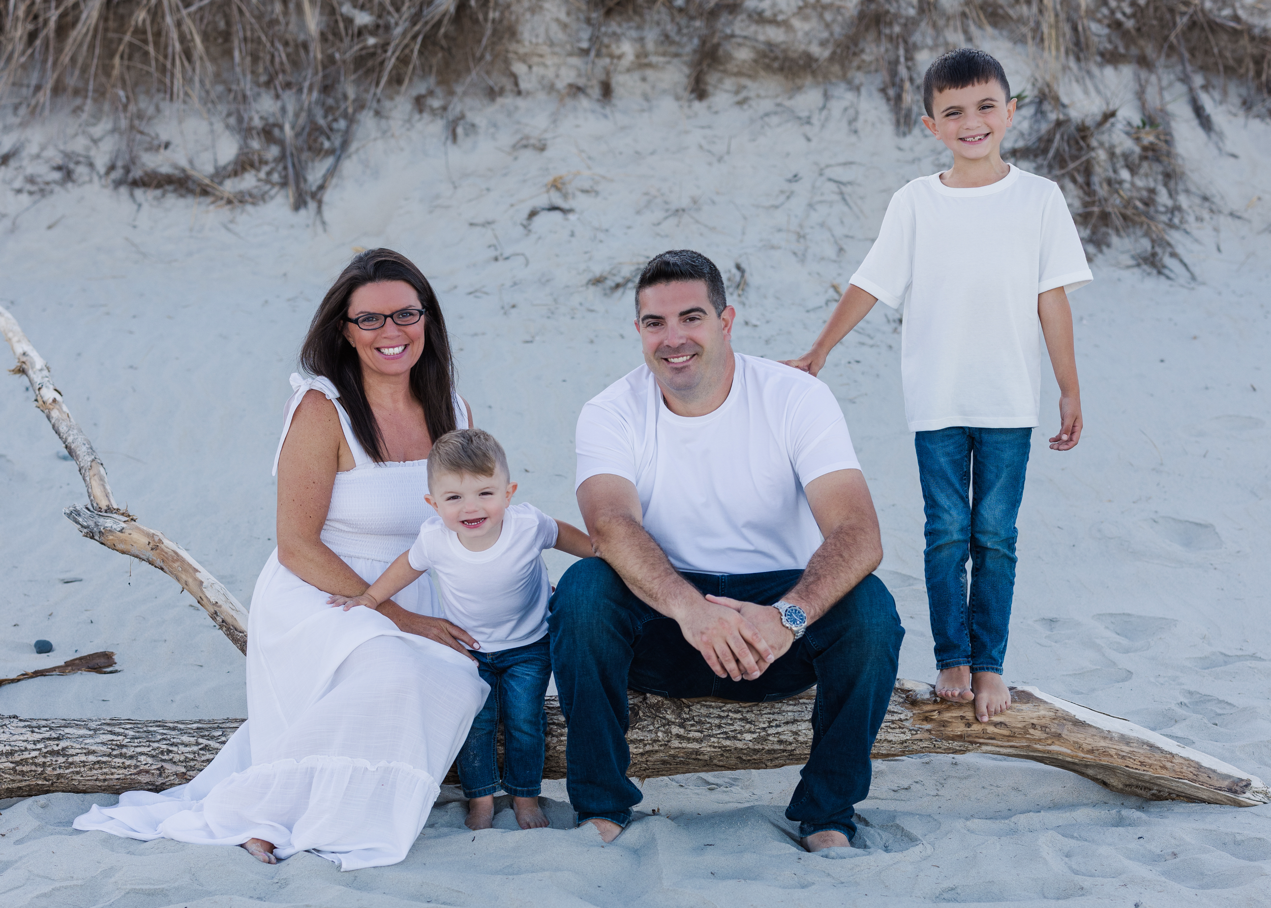 Family of five sitting on a driftwood log on the beach, smiling at the camera. The mother and two children are sitting on the left, with the father and another child standing on the right behind them, all dressed in white tops and blue jeans.