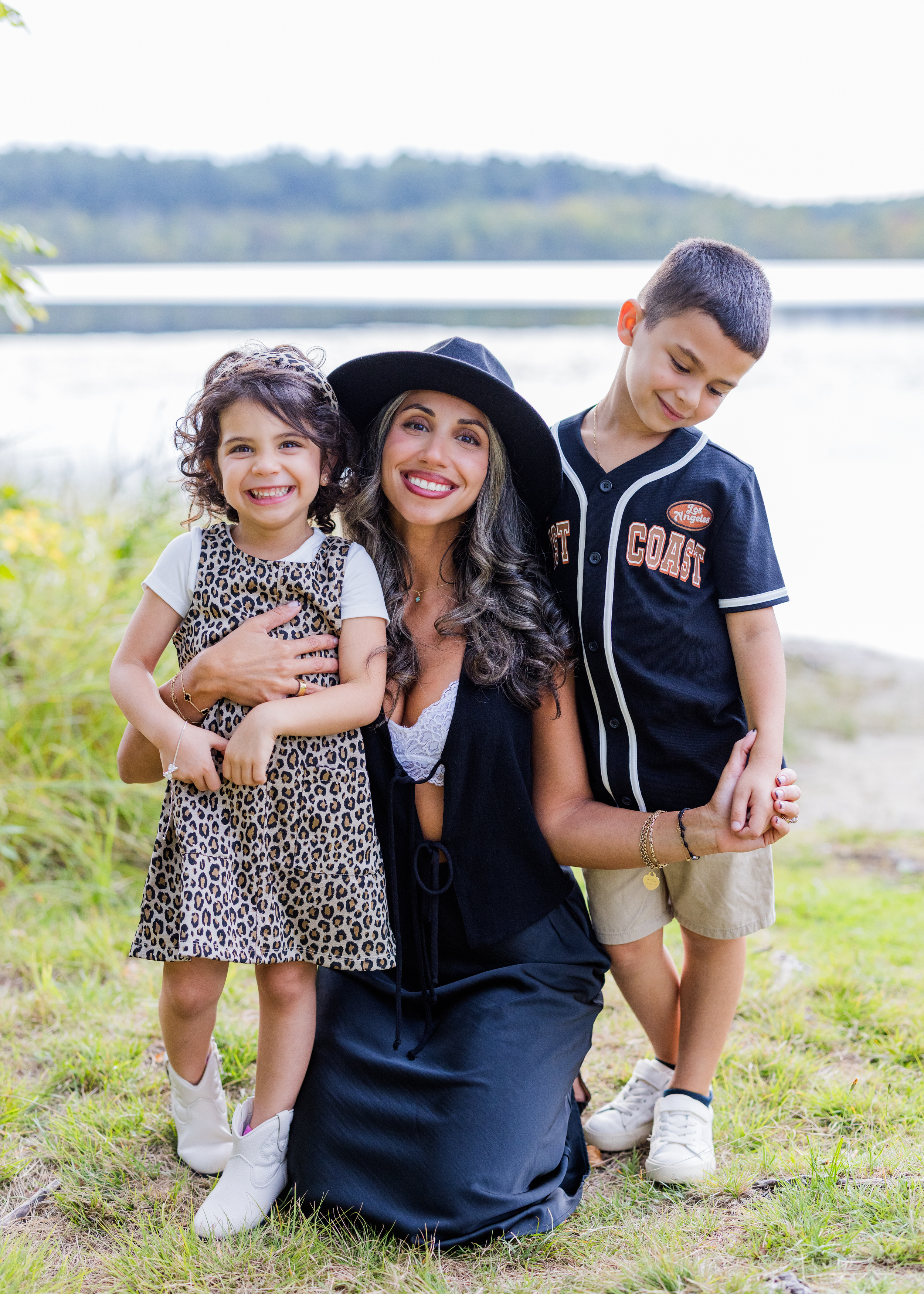 A woman with long dark hair wearing a black hat and dress, smiling, is posing outdoors with two children near a lake or river. The girl is wearing a leopard print dress, smiling, and has curly dark hair. The boy is wearing a black baseball jersey and shorts, looking down, and holding the woman's hand.