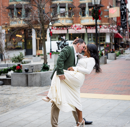 A couple dancing and smiling on a city sidewalk during winter with festive holiday decorations.