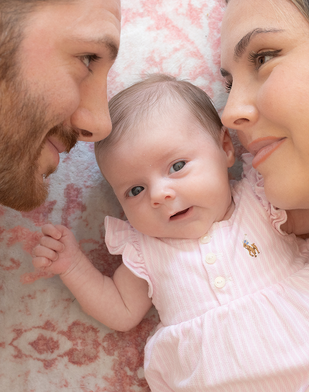 A smiling man and woman looking at a baby lying between them on a soft surface.