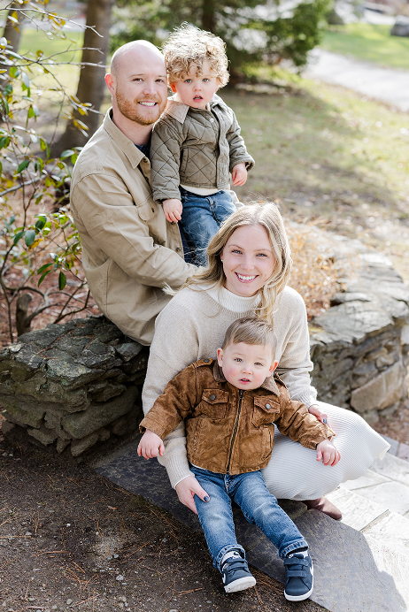 A family of four sitting outdoors on a stone ledge, with a father, mother, and two young children, smiling and enjoying a sunny day.