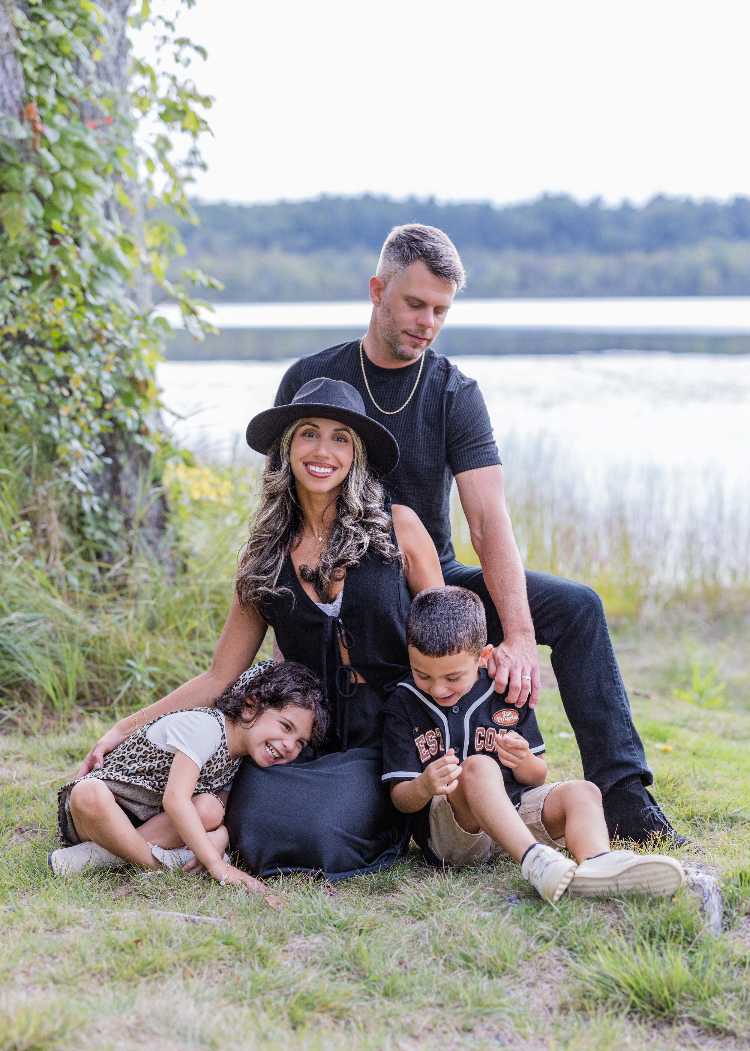 A family of five sitting on grass near a lake, smiling and enjoying nature with trees and water in the background.