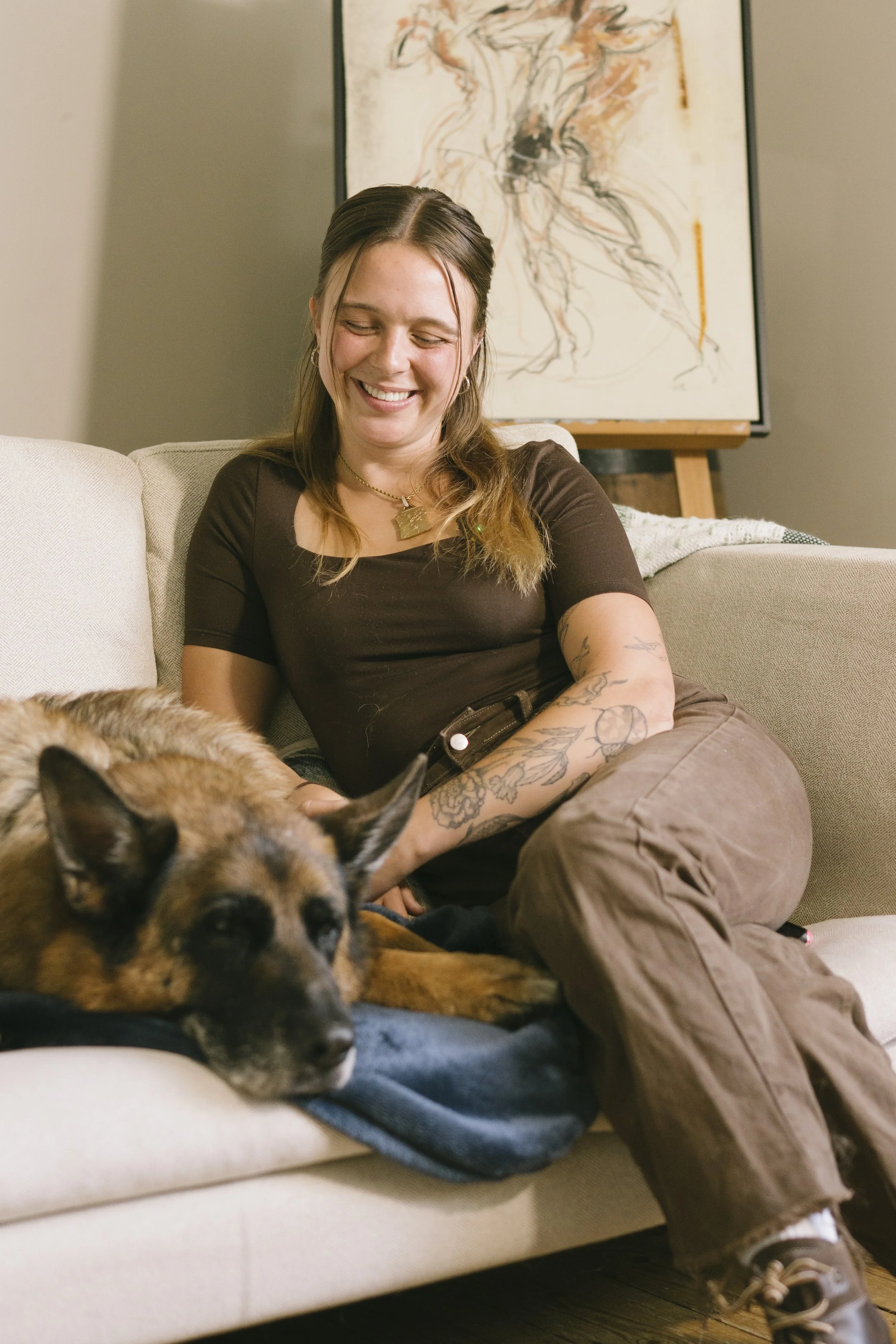 A woman with tattoos and brown hair smiling as she pets a sleeping dog on a beige sofa, with artwork hanging on the wall behind her.