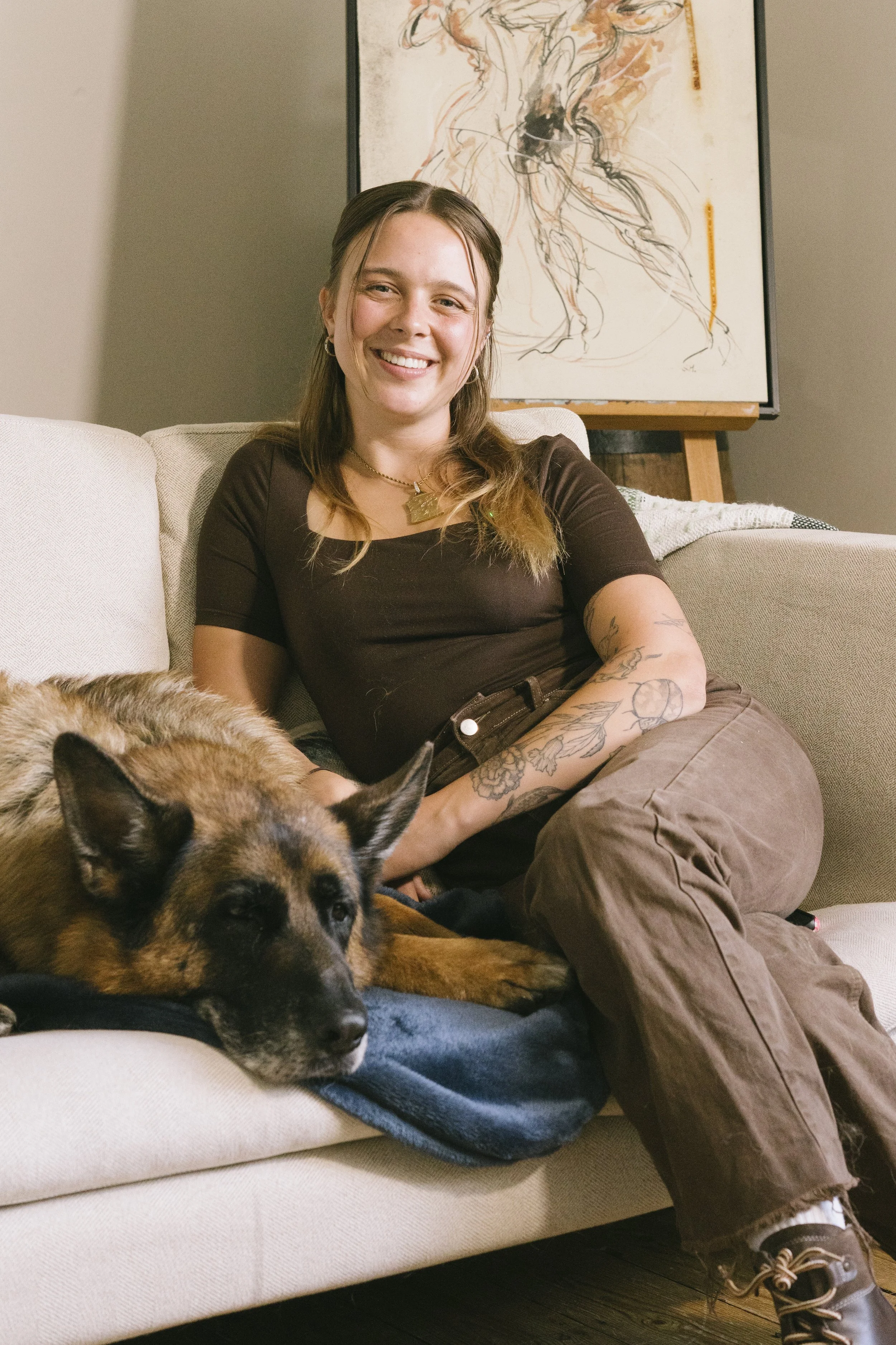 A young woman with tattoos sitting on a beige sofa, smiling at the camera, with a dog resting on a blanket in front of her. There is a colorful abstract painting behind her.