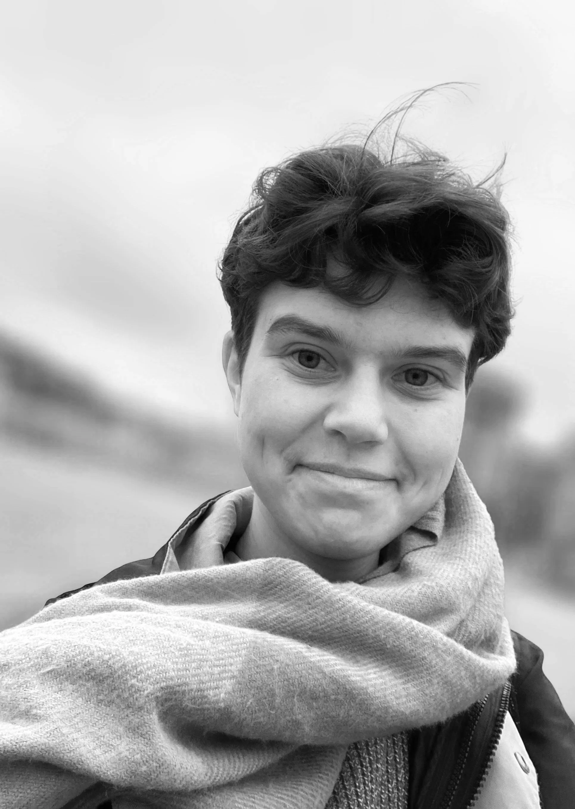 Un portrait en noir et blanc d'une jeune personne avec des cheveux courts et bouclés, portant un sweat ou une écharpe enroulée autour du cou, avec un léger sourire, dehors par temps nuageux.