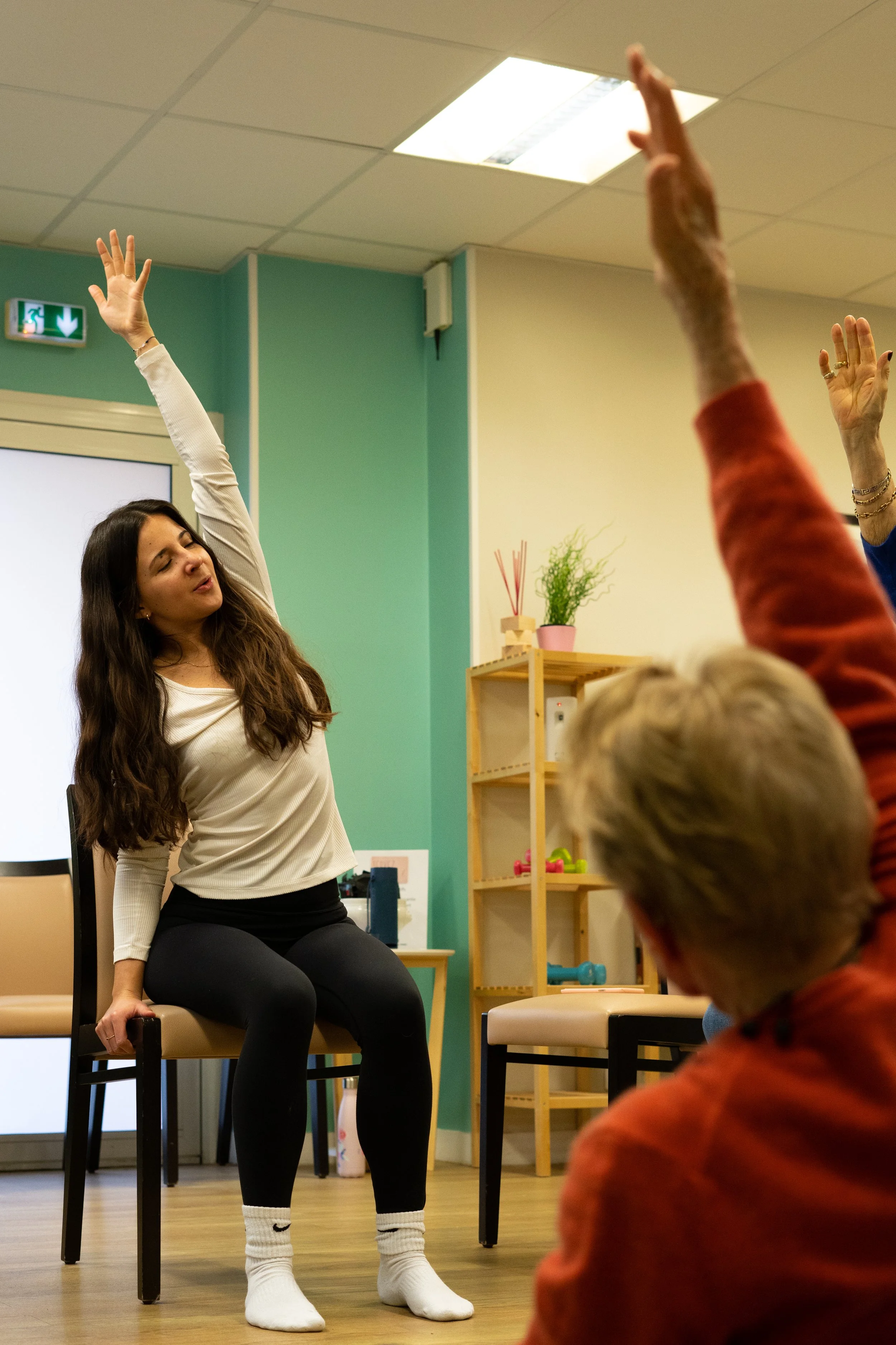 Une jeune femme assise sur une chaise effectue des étirements avec un bras levé, dans une salle de gym ou de yoga, en présence de plusieurs personnes, dont une avec la main levée.