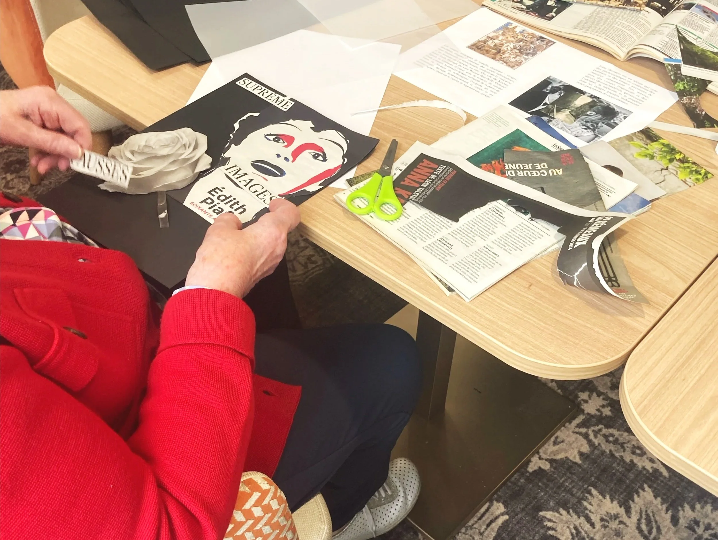 Une personne enroule une pièce de papier blanc à l'aide de ses mains, tandis qu'une autre personne, habillée en rouge, se trouve à côté. Sur la table en bois clair, il y a des ciseaux verts, des papiers déchirés, des magazine ouverts, et des feuilles de papier avec du texte, dans un environnement intérieur.