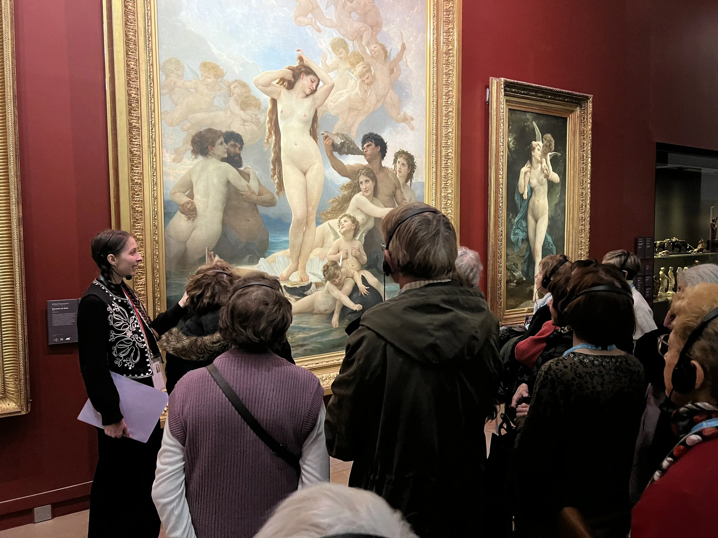 Un groupe de personnes écoute un guide touristique lors d'une visite de musée, observant un grand tableau classique représentant des figures mythologiques nues, entouré de cadres dorés.