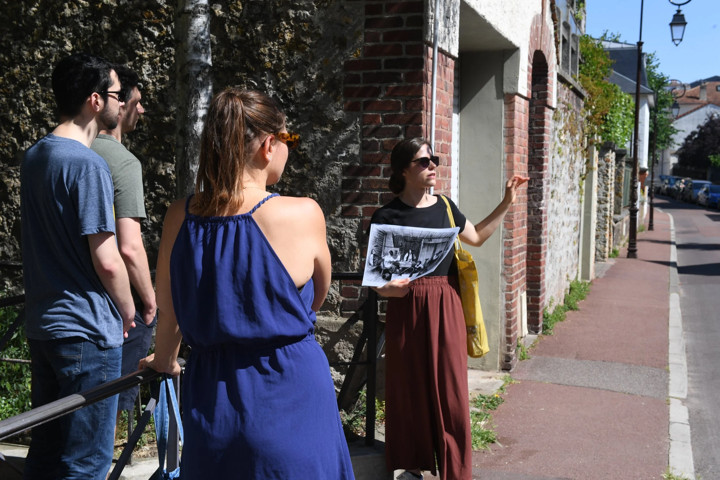 Une guide touristique explique un site à un groupe de touristes devant un mur en pierre, en plein air ensoleillé.
