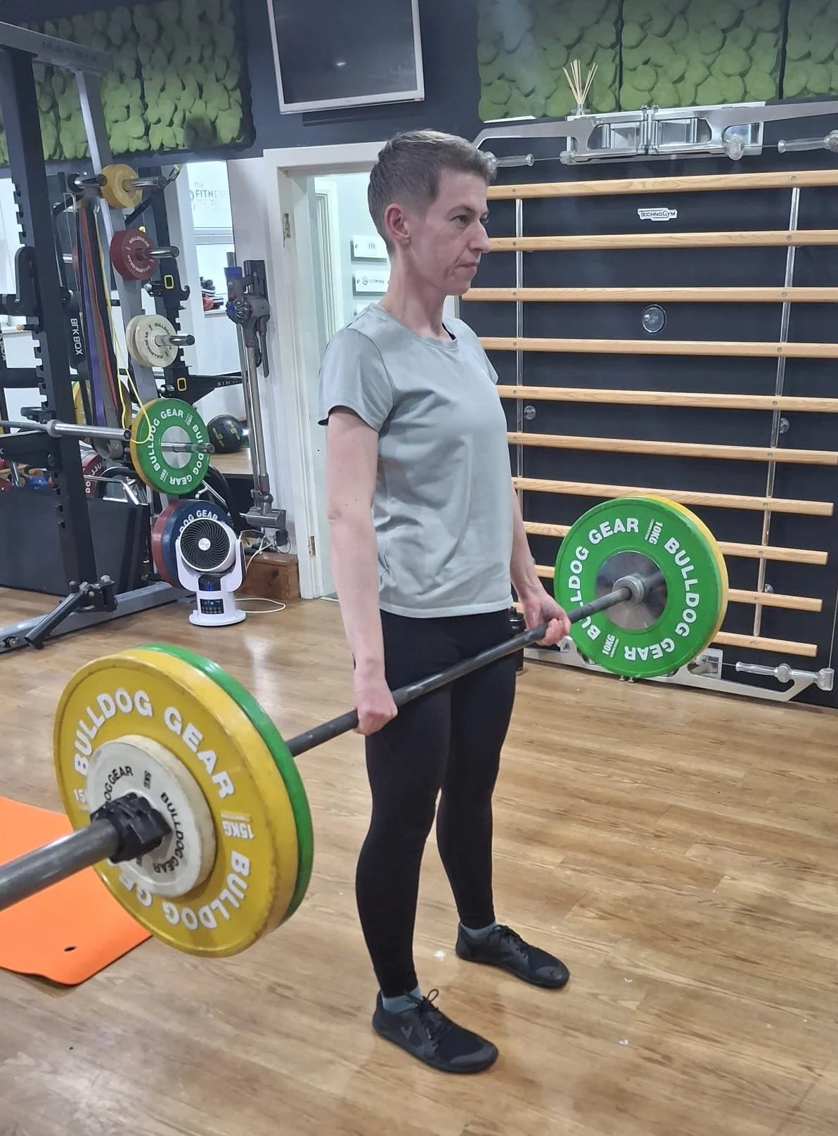 A woman lifting a barbell with yellow and green weight plates in a gym. She is standing on a wooden floor with gym equipment and wall bars in the background.