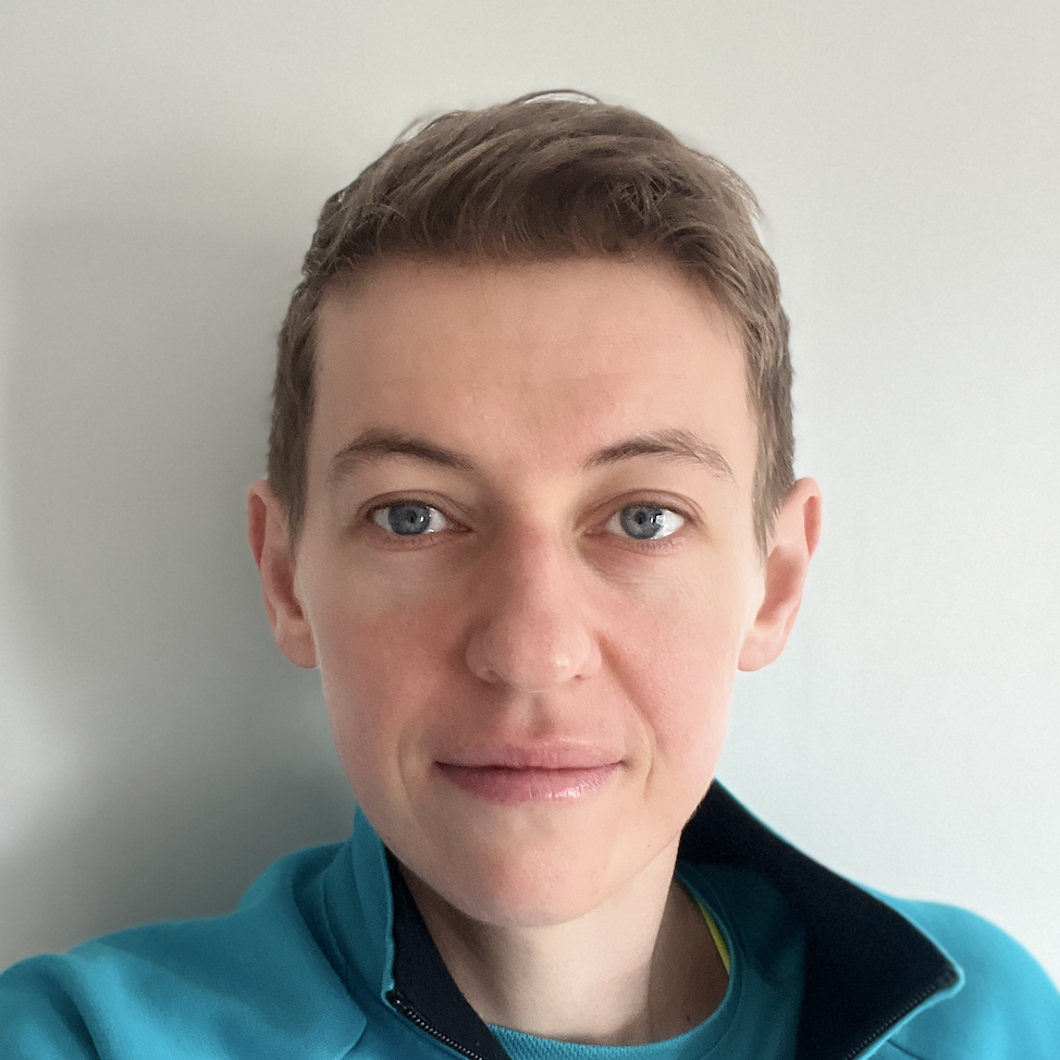 Close-up of a woman with short brown hair and blue eyes, wearing a blue athletic jacket, standing against a plain light background.