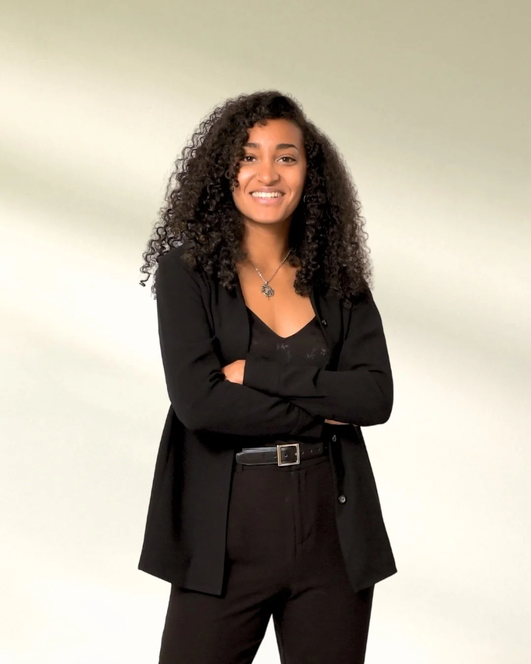 MacKenzie - a light-skinned African American woman with curly black hair and a bright smile, wearing a black blazer over a black camisole top, standing against a plain light-colored green-beige fade background.