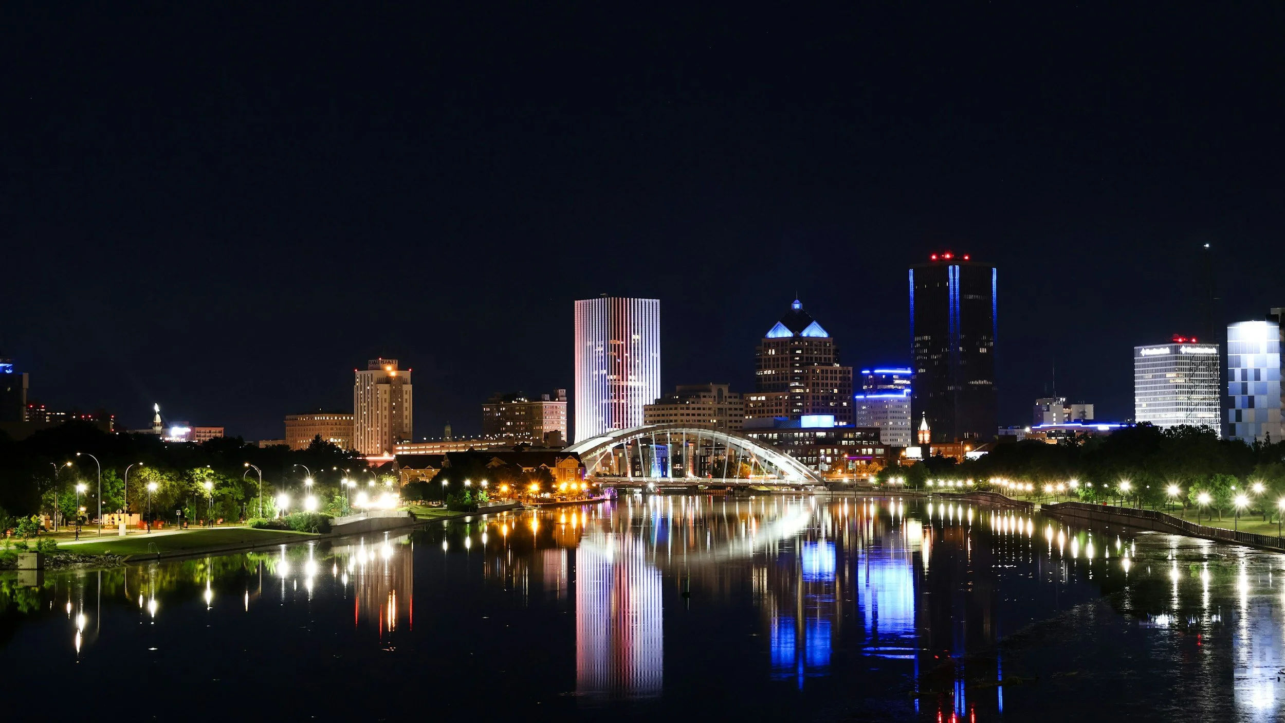 Nighttime city skyline with illuminated skyscrapers and their reflections in the water, including a bridge and a park with streetlights in the foreground.