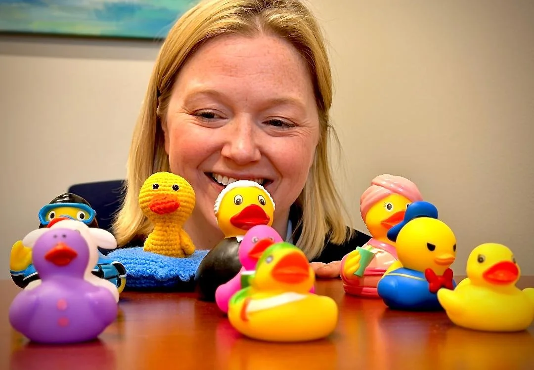 A smiling woman with blonde hair surrounded by colorful rubber duck toys on a wooden table.