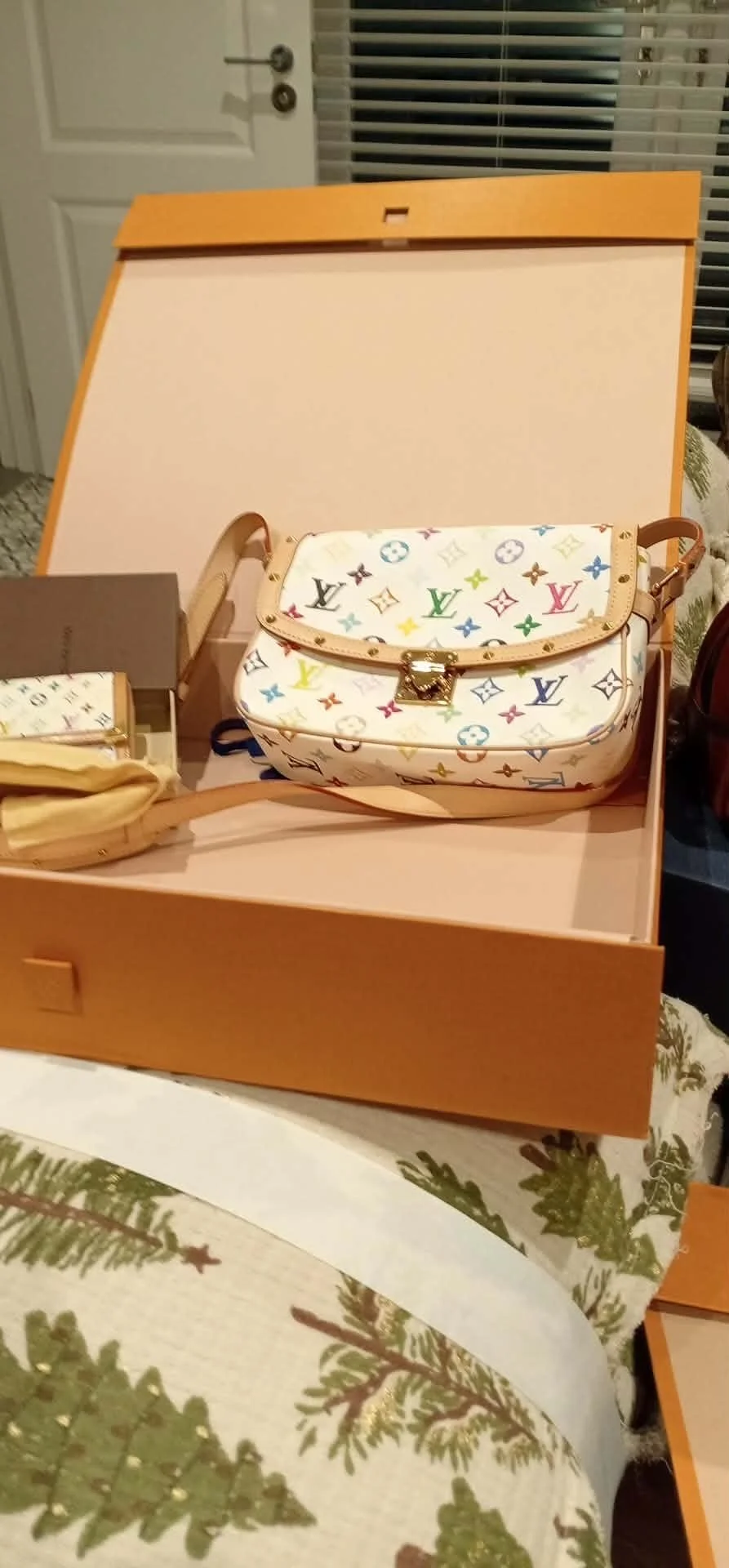 A multicolored Louis Vuitton handbag on an open pink box, with a smaller matching pouch nearby, on a table with Christmas-themed tablecloth.