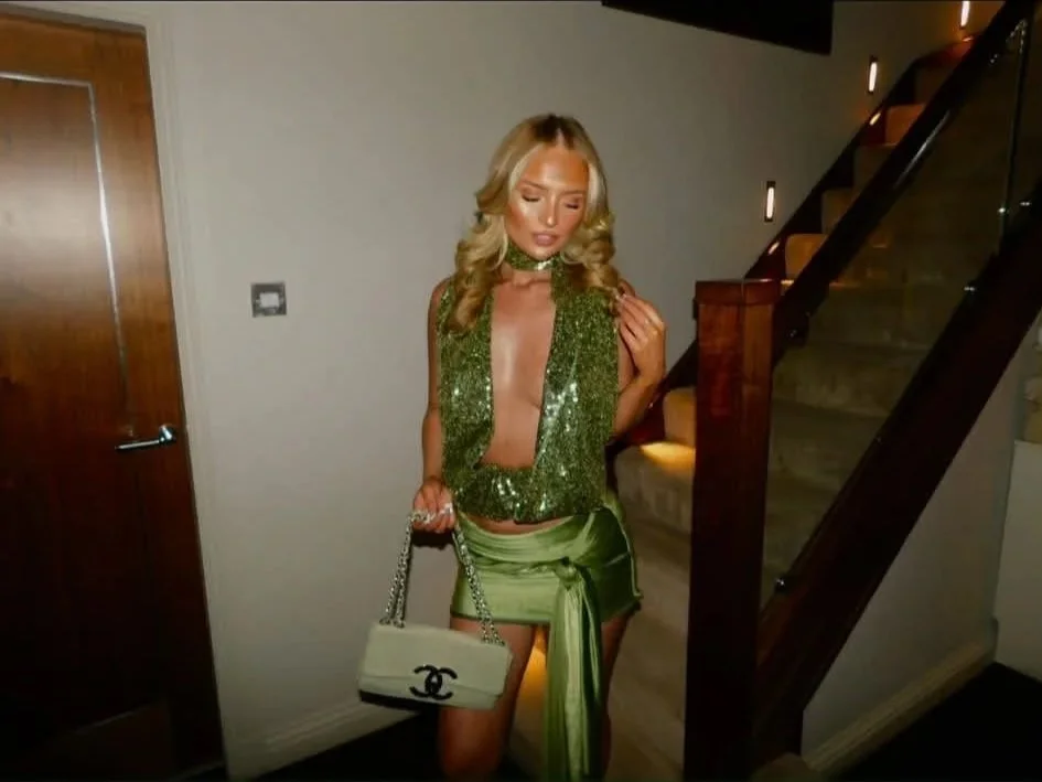 A woman with blonde hair, wearing a green sequined top, green satin skirt, and a Chanel purse, standing indoors near a staircase.