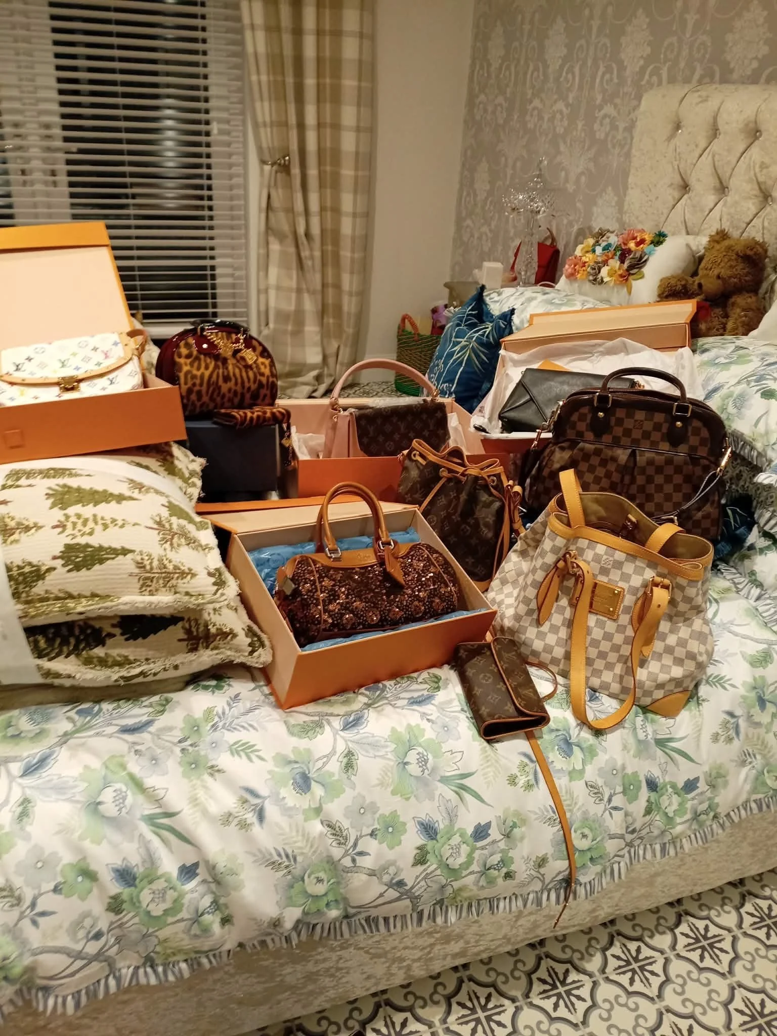 A bed with floral bedding and a variety of handbags and purses on top, some in boxes, in a room with patterned walls, curtains, and decorative pillows.
