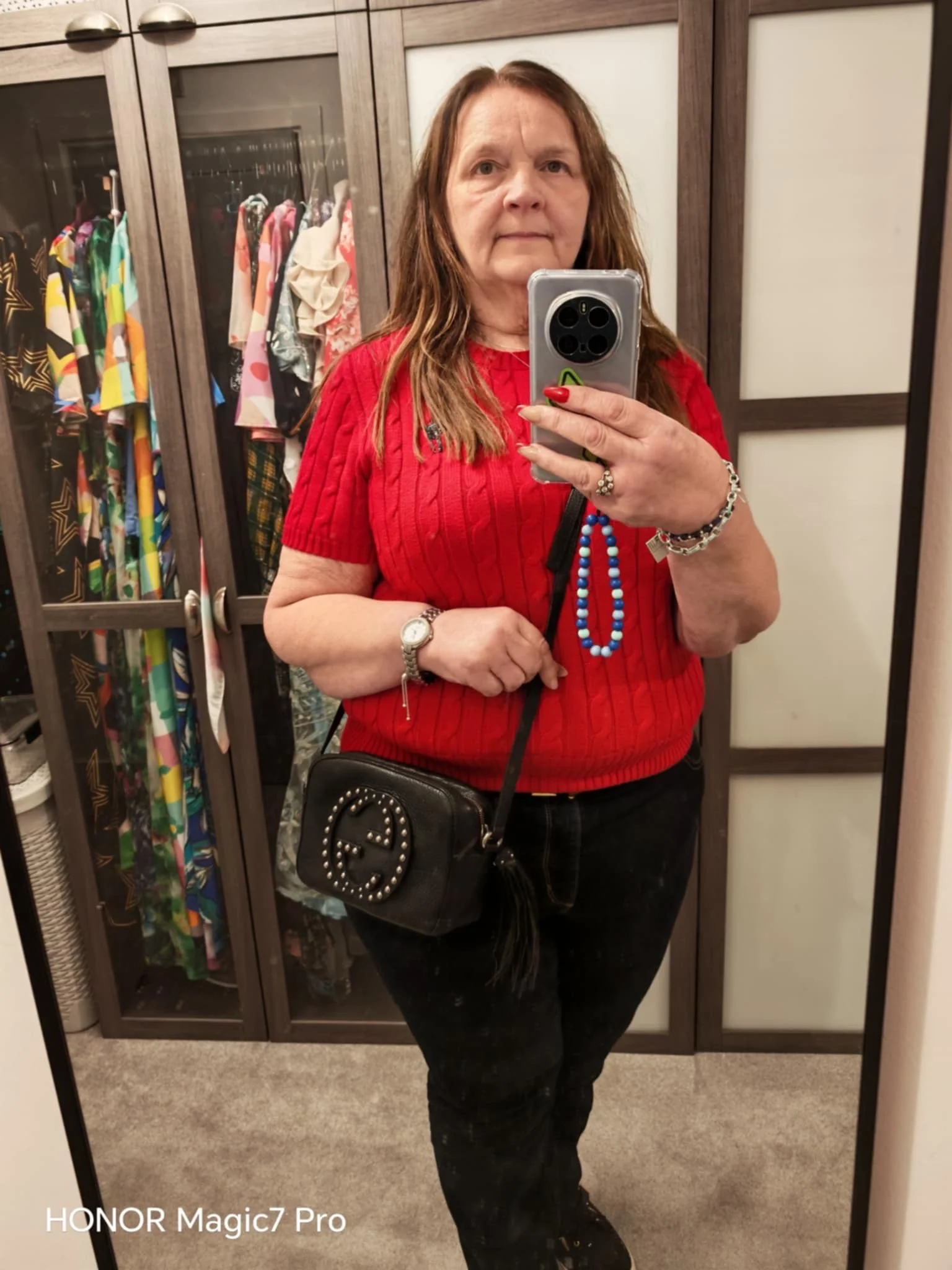 A woman taking a mirror selfie in a dressing room, wearing a red short-sleeve top, black pants, and carrying a black purse with a studded logo, accessorized with bracelets, a watch, and a necklace of blue beads. The dressing room has a wardrobe with 